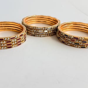 May include: Three sets of gold-toned bangles arranged in a triangular formation. Each set consists of multiple stacked rings featuring rectangular-cut stones in red, clear, and multicolored hues, accented with small white beads along the edges.