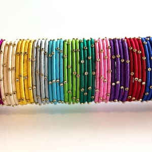 May include: A collection of colorful bangles in a rainbow of hues, including pink, yellow, green, and blue. Each bangle is adorned with small, gold-colored embellishments. The bangles are stacked together, creating a vibrant display.