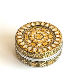 May include: A round, decorative box with a detailed floral design. The lid and sides feature gold and white patterns on a dark background. The box has a clear, cylindrical base, and the lid is adorned with a central floral motif.