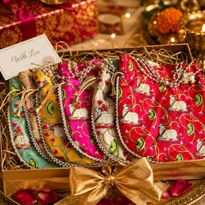 May include: A gift box containing five drawstring pouches in teal, peach, yellow, magenta, and red, each featuring a repeating pattern of white cows and lotus flowers. The pouches are adorned with pearl-like beaded strings and a card reading With Love.