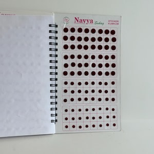 May include: Open notebook with blank white pages next to a sheet of burgundy dot stickers. The sticker sheet is labeled "Navya Suhag Sticker Kumkum". The notebook has a black spiral binding.