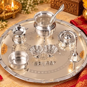 May include: A polished silver-toned metal puja thali set featuring a large round tray with a decorative border, a central kalash with a spoon, a small diya, a bell, two small bowls, and a central incense holder, all arranged on a reflective surface.