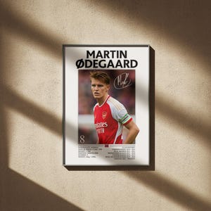 May include: Framed poster featuring Martin Ødegaard in an Arsenal jersey. The red and white jersey has the Emirates logo. The poster includes the player's name, signature, and career stats. The background is a neutral wall with shadows.