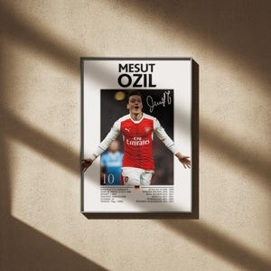 May include: Framed poster featuring Mesut Özil in an Arsenal jersey, arms outstretched. The red and white jersey has the Emirates logo. The poster includes the player's name, signature, and career highlights. The background is a neutral beige.