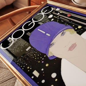 May include: A framed print featuring a stylized illustration of a person wearing a purple cloche hat and a pearl necklace. The background depicts a dark cityscape at night with illuminated windows. The text "VOGUE" is printed in large white letters.