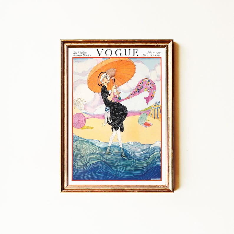 May include: A framed vintage Vogue magazine cover from July 1, 1919, featuring an illustration of a figure in a black dress holding an orange parasol while standing in blue ocean waves on a beach with a pink circle and a striped tent in the background.