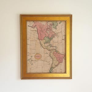 May include: A framed vintage-style map of North and South America featuring a puzzle-piece texture. The map displays landmasses in muted pink and green tones against a beige background, enclosed in a wide, metallic gold-toned rectangular frame.