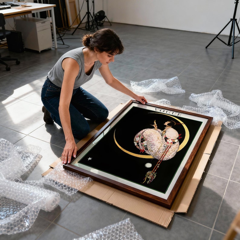 以下が含まれることがあります： 黒い背景に三日月の中の女性を描いた、額装された「VOGUE」アートプリント。 プリントは茶色の木製フレームで、スタジオ環境で人が扱っています。 近くにはプチプチがあります。
