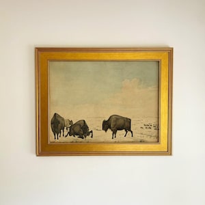 May include: A framed print featuring a landscape illustration of several bison in a vast, open field under a pale sky. The artwork is enclosed in a wide, metallic gold frame with a smooth, polished finish and a rectangular border design.