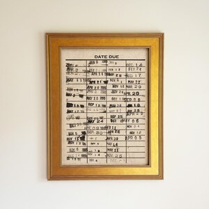 May include: A rectangular gold-toned frame containing a vintage library date due card. The card features a grid with black horizontal and vertical lines, printed with the text DATE DUE at the top, and filled with various stamped dates in black ink.