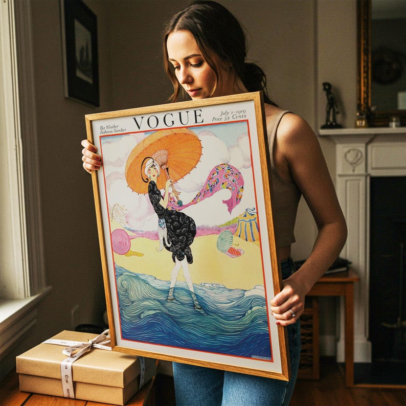 Puede incluir: Una impresi&oacute;n enmarcada de la portada de la revista Vogue del 1 de julio de 1919, con un precio de 35 centavos. La ilustraci&oacute;n muestra a una persona con vestido negro sosteniendo una sombrilla naranja, de pie en olas azules con una bufanda rosa y una tienda amarilla.