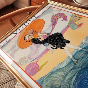 Puede incluir: Una portada de revista Vogue vintage enmarcada del 1 de julio de 1919, titulada Hot Weather Fashions Number. La ilustraci&oacute;n muestra a una figura con vestido negro sosteniendo una sombrilla naranja, caminando en una playa junto a olas azules.
