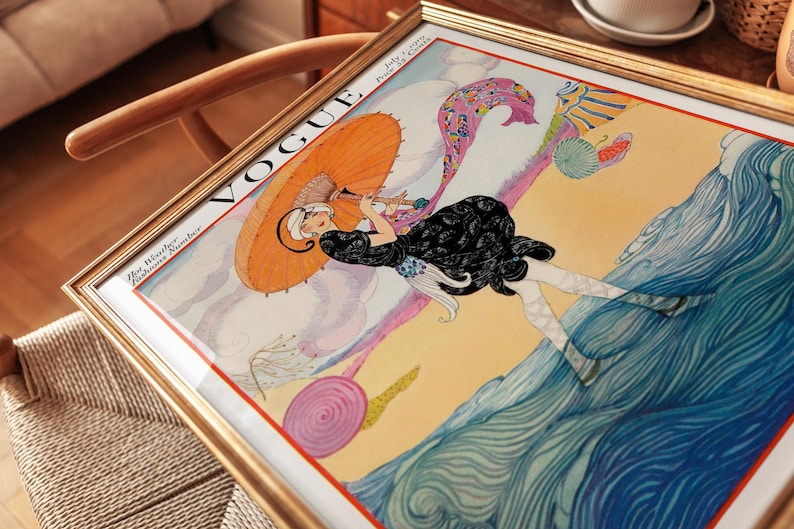 Puede incluir: Una portada de revista Vogue vintage enmarcada del 1 de julio de 1919, titulada Hot Weather Fashions Number. La ilustraci&oacute;n muestra a una figura con vestido negro sosteniendo una sombrilla naranja, caminando en una playa junto a olas azules.