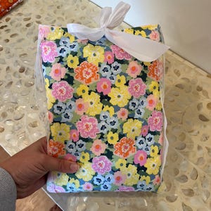 May include: A floral fabric item, wrapped and tied with a white ribbon. The fabric features a colorful pattern of pink, yellow, orange, and blue flowers on a dark green background. The edges are trimmed with white lace.
