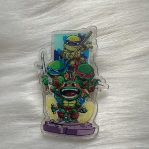 May include: A clear acrylic stand featuring the Teenage Mutant Ninja Turtles. The stand depicts the four turtles in their signature colours, with weapons in hand, set against a cityscape backdrop. The base is purple.