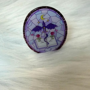 May include: A circular pin featuring a stained-glass style design in shades of purple and blue. The illustration depicts a purple dragon perched on a sword embedded in an open book, flanked by two red roses, a yellow crescent moon, and small hearts.