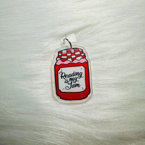 May include: A clear acrylic keyring shaped like a jam jar, featuring a red and white checked lid. The jar is filled with pink jam and has the text "Reading is my Jam" in white script.