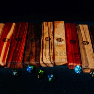 May include: A collection of wooden dice boxes in various wood grains and colors, including light, red, and dark brown. Each box has a logo and is rectangular. Several multi-sided dice are scattered in front of the boxes.