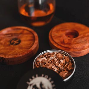 Puede incluir: Se muestran dos posavasos de madera y una lata de virutas de madera. Los posavasos son circulares, con un color marrón intenso y un diseño tallado. La lata está abierta, revelando las virutas de madera. Un vaso de líquido ámbar está en el fondo.