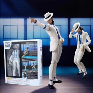 May include: Two articulated Michael Jackson action figures wearing white suits, white fedoras, and blue shirts. One figure points forward while the other leans. A rectangular product box with text and images of the figure is positioned on the left.
