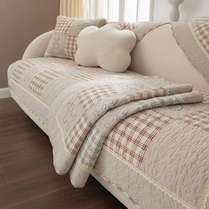 May include: A beige sofa with a patchwork quilt and matching cushions. The quilt features a plaid pattern in red, white, and beige squares, with embroidered bunny figures. A cloud-shaped cushion and a square cushion with a bunny design are also visible.