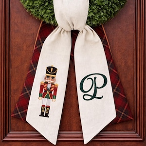 May include: A decorative fabric ribbon hanging from a green wreath, featuring a cream-coloured front with a printed nutcracker figure on one side and a dark green letter P on the other, backed by a red and dark green plaid fabric pattern.