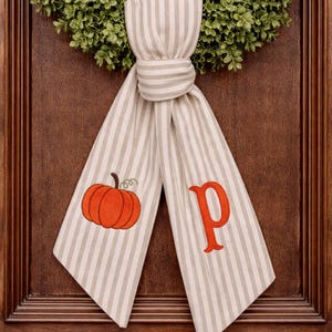 Puede incluir: Un colgador de puerta de tela a rayas beige y blancas con una calabaza naranja bordada y la letra "P". La tela está anudada y cuelga frente a una corona verde.