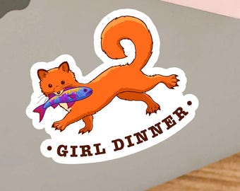 May include: A die-cut sticker featuring an orange fox running while holding a colorful, patterned fish in its mouth. Below the fox, the text "GIRL DINNER" is printed in brown, capital letters, flanked by two small brown dots on either side.