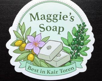 May include: A circular sticker featuring the text Maggie's Soap and Best in Kair Toren. The design includes two light-colored rectangular soap bars with shell icons, green olive branches with yellow olives, a purple flower, and green berries.