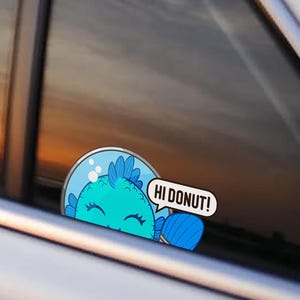 May include: A circular sticker applied to a car window featuring a cartoon blue fish inside a glass bowl. The fish has a smiling expression with closed eyes and a speech bubble extending to the right that contains the text HI DONUT!