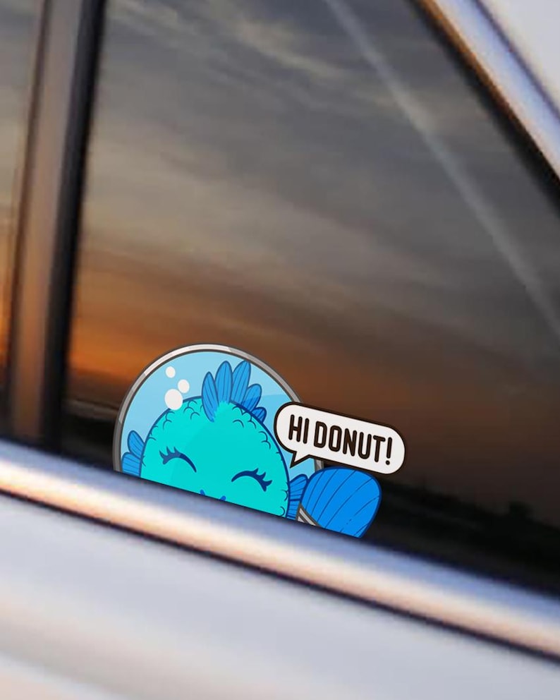 May include: A circular sticker applied to a car window featuring a cartoon blue fish inside a glass bowl. The fish has a smiling expression with closed eyes and a speech bubble extending to the right that contains the text HI DONUT!