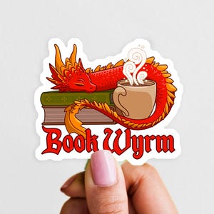 May include: A die-cut sticker featuring a red dragon with orange accents resting on a stack of two books next to a steaming mug of coffee. The text "Book Wyrm" is printed in a bold red gothic-style font at the bottom of the illustration.