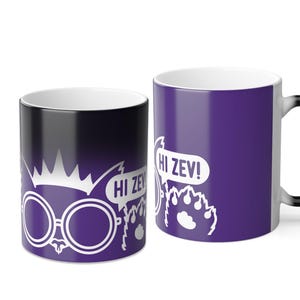 May include: Two ceramic mugs with a white interior. One mug is black with a white illustration of glasses, a crown, and the text "HI ZEV!". The other mug is purple with a white paw print and the text "HI ZEV!".