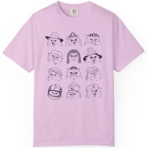 May include: A light purple short-sleeve t-shirt featuring a dark purple graphic print of twelve cats wearing various hats and accessories, including crowns, a wizard hat, a motorcycle helmet, and a sailor cap, arranged in a grid pattern.