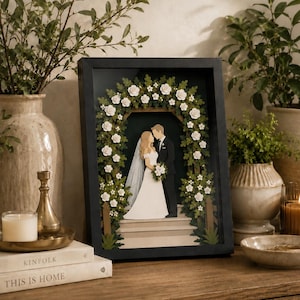 May include: A framed paper art wedding scene. A bride and groom stand under a floral archway. The artwork is in a black frame and is displayed on a wooden surface, surrounded by decorative items, such as vases and candles.