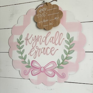 May include: Pink and white gingham baby door hanger with a green wreath and a pink bow. The hanger has the name "Kyndall Grace" written in cursive. A separate wooden tag is attached with the words "date", "time", "lbs", and "in."