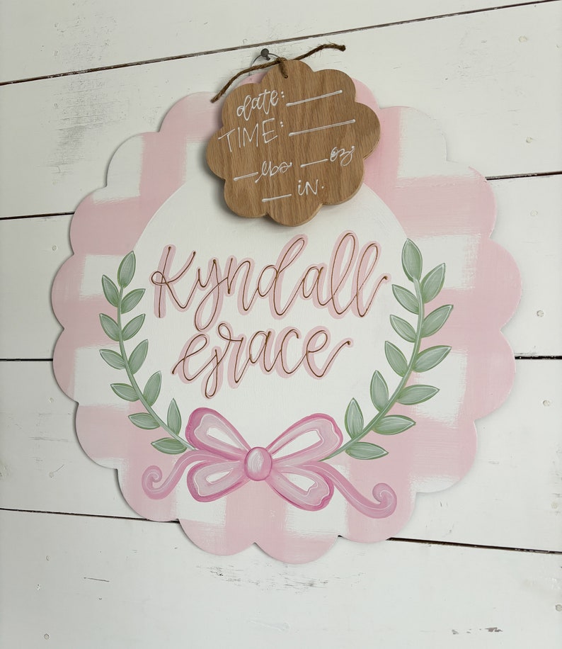 May include: Pink and white gingham baby door hanger with a green wreath and a pink bow. The hanger has the name "Kyndall Grace" written in cursive. A separate wooden tag is attached with the words "date", "time", "lbs", and "in."