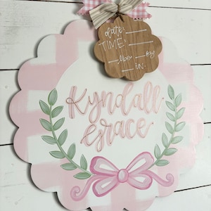 May include: Pink and white gingham baby door hanger with a green wreath and a pink bow. The name "Kyndall Grace" is written in gold script. A wooden tag with the words "date", "time", "lbs", and "in." is attached to the top.