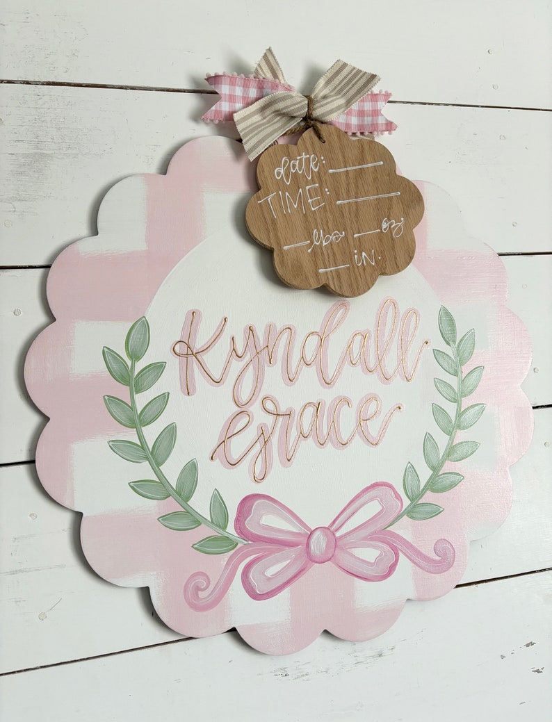 May include: Pink and white gingham baby door hanger with a green wreath and a pink bow. The name "Kyndall Grace" is written in gold script. A wooden tag with the words "date", "time", "lbs", and "in." is attached to the top.