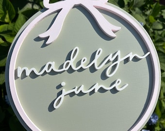 May include: A round, sage green wooden sign with a white bow at the top. The sign has a white border and features the names "madelyn june" in a cursive font. The sign is held in front of a background of green leaves and blue flowers.