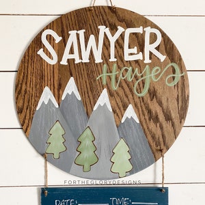 May include: A round wooden sign with a mountain and pine tree design. The sign is painted in shades of brown, gray, and green. The name "Sawyer Hayes" is written in white and green paint. A smaller chalkboard sign hangs below with the words "Date", "Time", "Weight", and "Length" written in white paint.