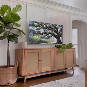 May include: A light wood cabinet with four doors and woven cane panels. A large TV displays a painting of a tree with hanging beads. A potted plant and a woven basket are also in the room. The room has a neutral color palette.