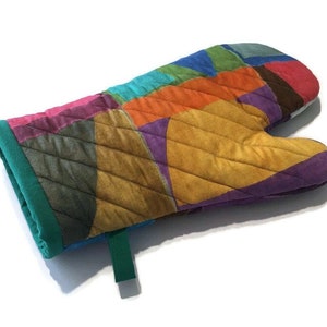 May include: A colourful quilted oven glove with a patchwork design. The glove features a variety of colours, including pink, orange, yellow, purple, and green. It has a green cuff and a green hanging loop. Designed to protect hands from heat.