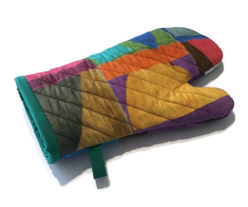 May include: A colourful quilted oven glove with a patchwork design. The glove features a variety of colours, including pink, orange, yellow, purple, and green. It has a green cuff and a green hanging loop. Designed to protect hands from heat.