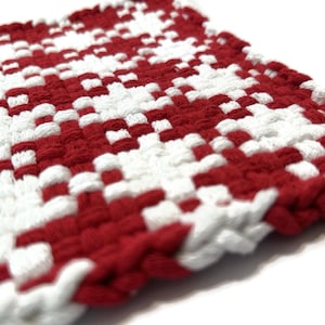 May include: Close-up of a square, woven trivet in a red and white checkerboard pattern. The trivet is made of thick, textured material, with the red and white threads interlaced to create the design. The edges are finished with a thicker, braided border.