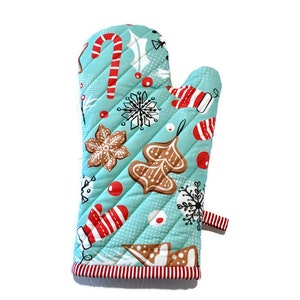 May include: A quilted oven mitt with a light blue background and a repeating Christmas-themed pattern. The pattern includes gingerbread cookies, candy canes, snowflakes, and red accents. The mitt has a red and white striped trim and a small loop.