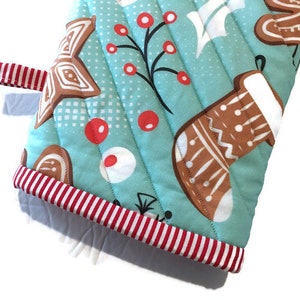 May include: A quilted oven mitt with a light blue background and a repeating pattern of gingerbread cookies, red berries, and white accents. The mitt has a red and white striped trim and a red and white striped loop.