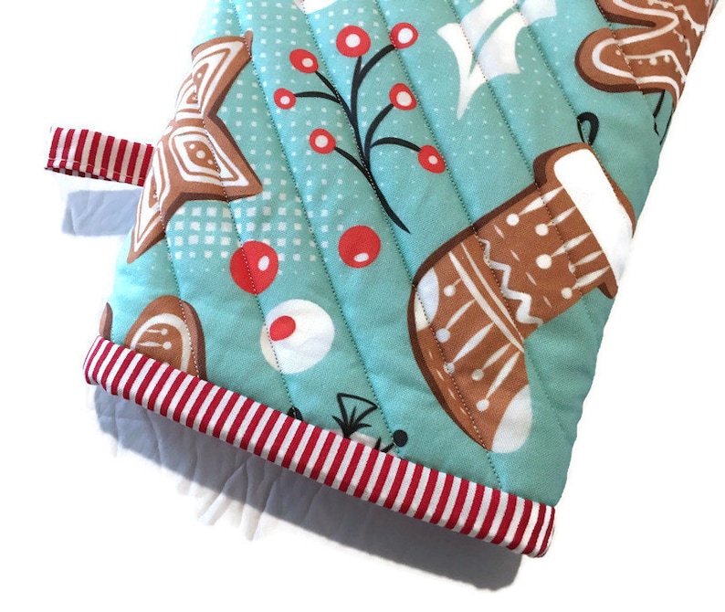 May include: A quilted oven mitt with a light blue background and a repeating pattern of gingerbread cookies, red berries, and white accents. The mitt has a red and white striped trim and a red and white striped loop.