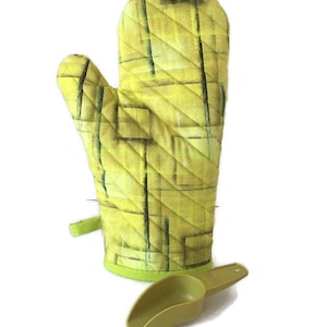 May include: A yellow quilted oven mitt with a green trim and a matching scoop. The mitt has a geometric pattern in shades of yellow and black. The scoop is olive green.