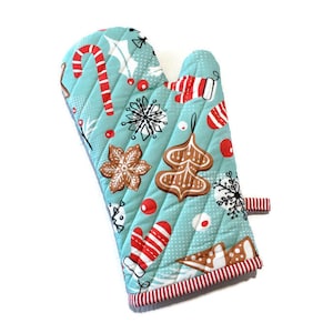 May include: A quilted oven mitt with a light blue background and a repeating Christmas-themed pattern. The pattern includes red and white candy canes, gingerbread cookies, snowflakes, and red berries. The mitt has a red and white striped trim.
